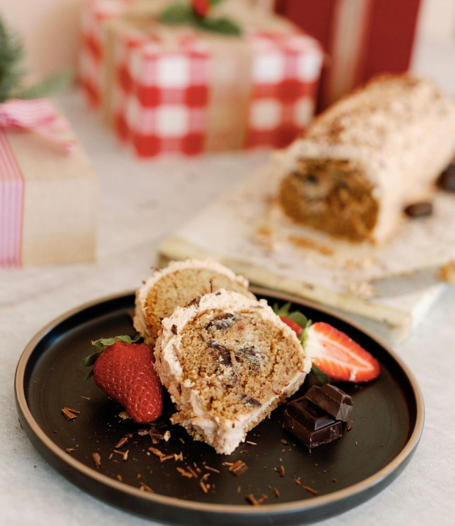 Two slicers of gluten free Christmas log on a brown plate. chrsitams log in background with red and white checked wrapped parcel