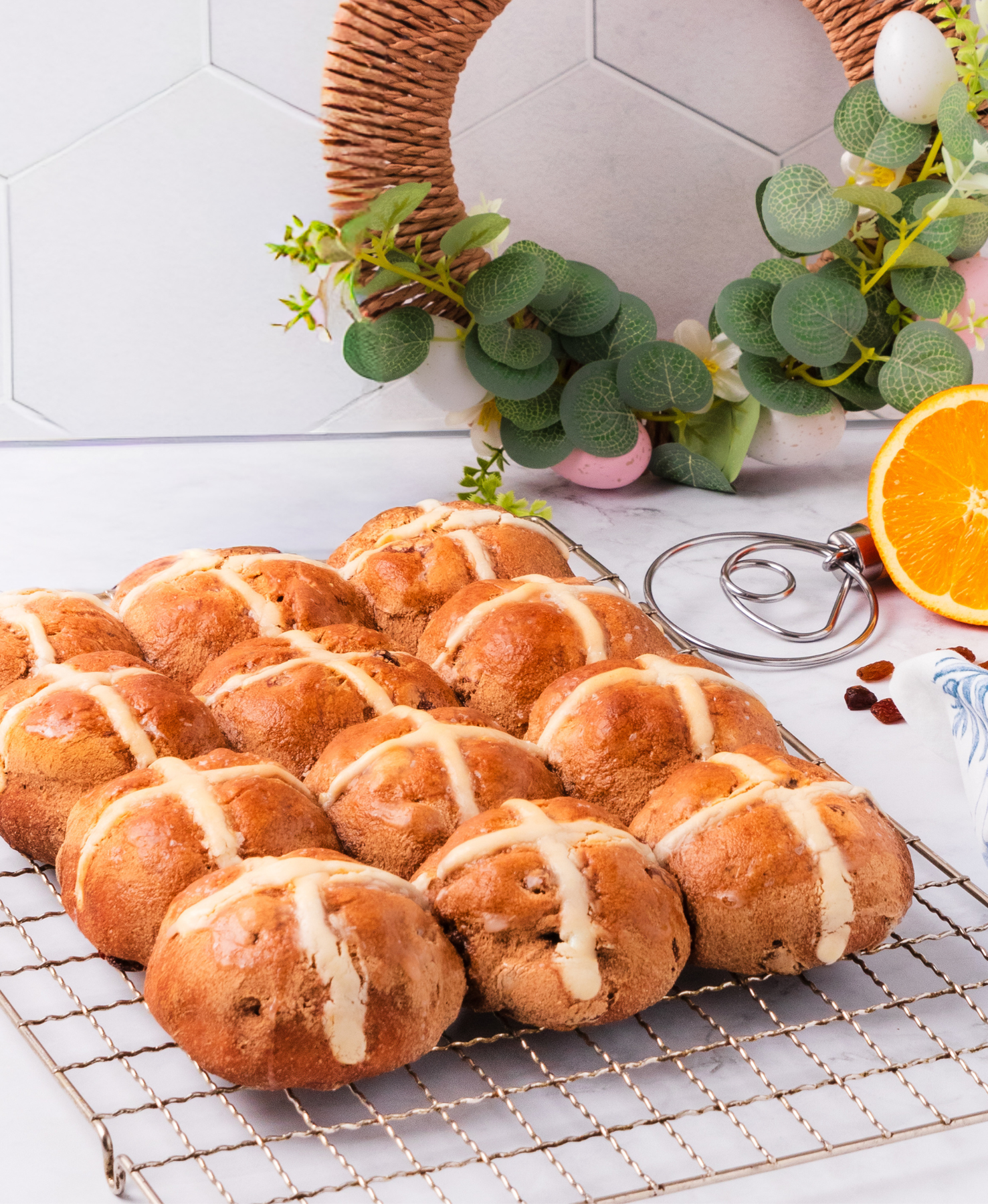 Gluten free hot cross buns on cooling rack with half an orange and a whisk and easter decorations in the background 