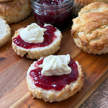 Load image into Gallery viewer, Scones with jam and cream on a wooden board with a jar of jam and a spoon.