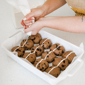 Lady piping crosses onto gluten free hot cross buns. 12 buns in a white dish  on a stone bench. Baker has white nail polish  