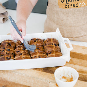Brushing glaze onto gluten free hot cross buns with a grey silicon pastry brush. Dish is sitting on wooden board. Glaze is in a white ramekin in front of buns  