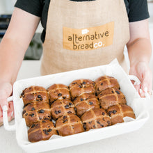 Load image into Gallery viewer, Holding white baking dish with 12 gluten free hot cross buns. Lady is wearing hessian apron with the words Alternative Bread co on it.