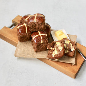 Gluten Free hot cross buns on wooden board with butter in white dish. One bun cut in half spread with butter 